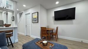 A modern living room with a sofa, ottoman, and a dining area with a table and chairs. A television is mounted on the wall, and there is a staircase leading to another level. - Heart of the Hamptons, Waterfront with Pool!  1 Bed/1 Bath Townhome (Hampton Bays)