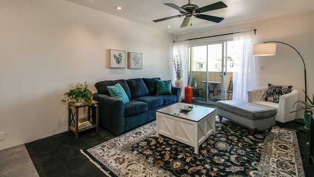 Un salon avec un canapé bleu foncé, une table basse blanche, un pouf gris, une lampe sur pied et un ventilateur de plafond.
