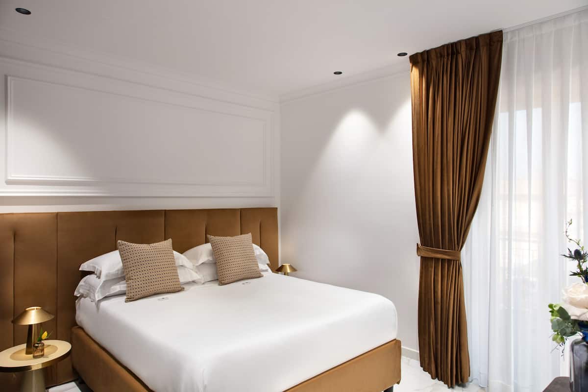 Une chambre d’hôtel moderne avec un grand lit, une table de chevet ronde surmontée d’une lampe, et une vue sur la ville à travers de fines rideaux.