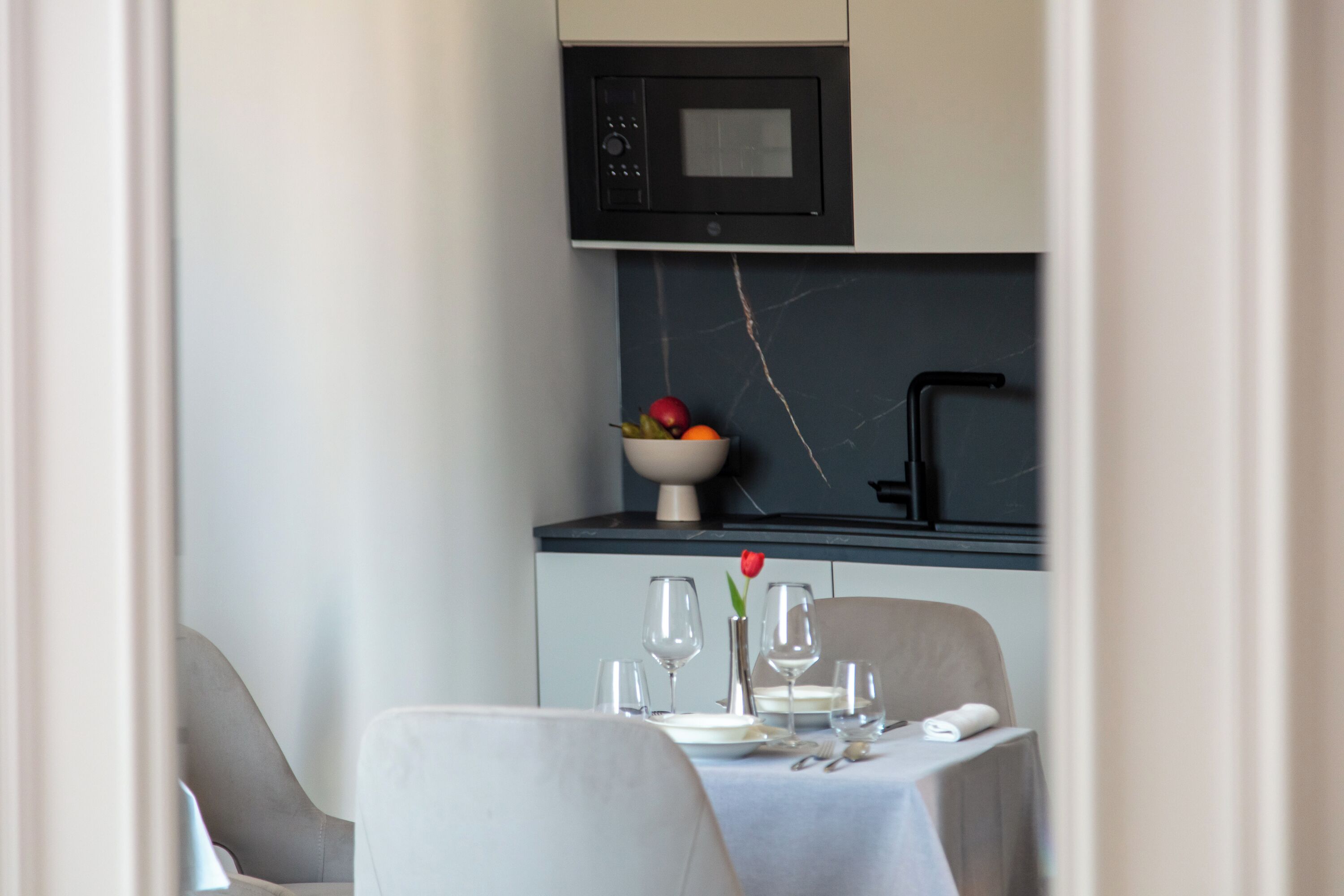 A dining area with a table set for two, a bowl of fruit, and a vase with a single red rose.