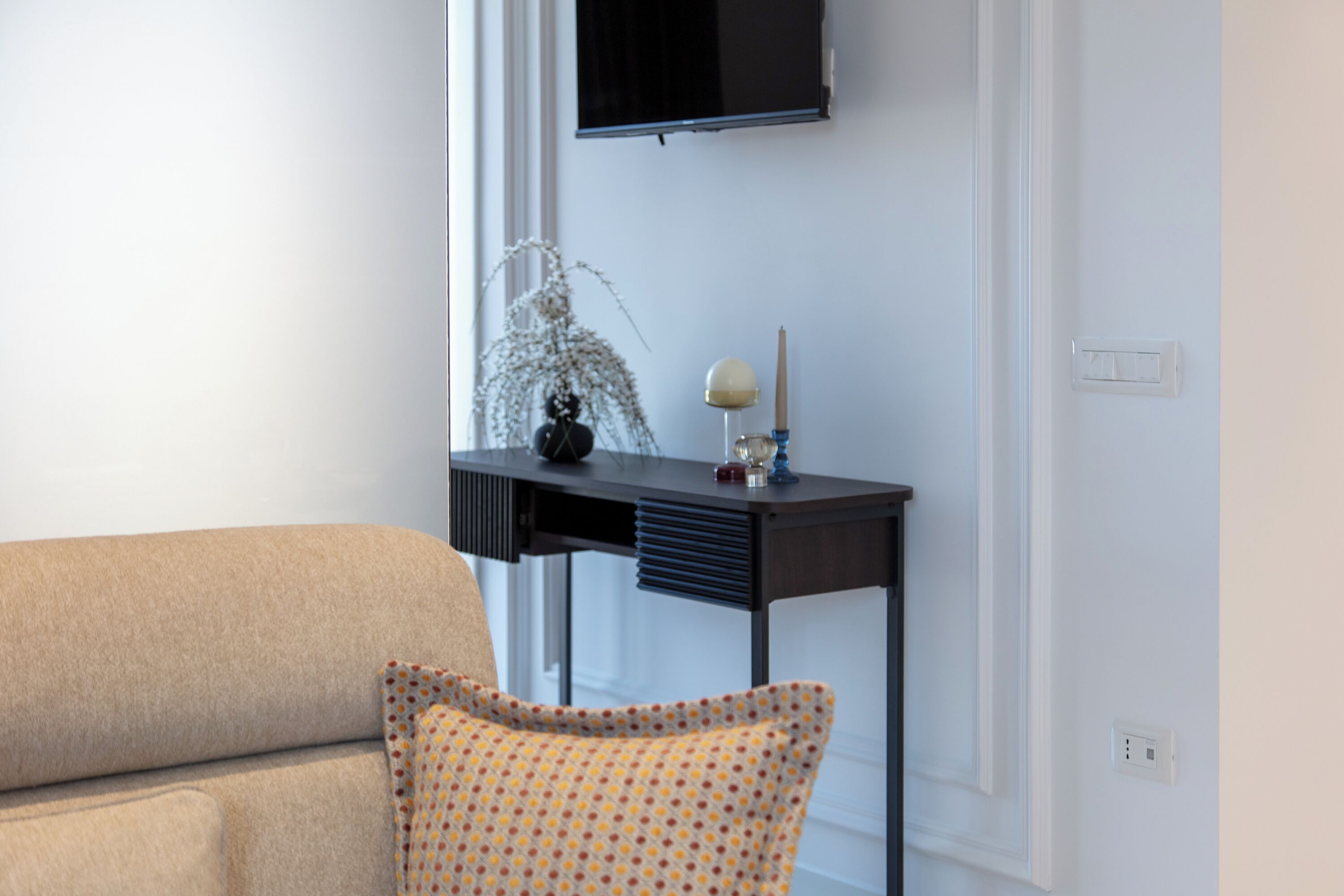Una sala moderna con un sofá color beige, un sillón estampado, una mesita de noche de madera oscura y un televisor montado en la pared.