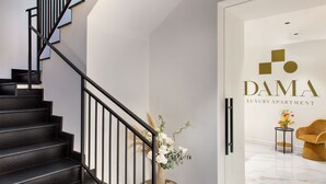 Een moderne trap met zwarte leuningen leidt naar een kamer met een witte muur en een bord waarop 'DAMA LUXURY APARTMENT' staat.