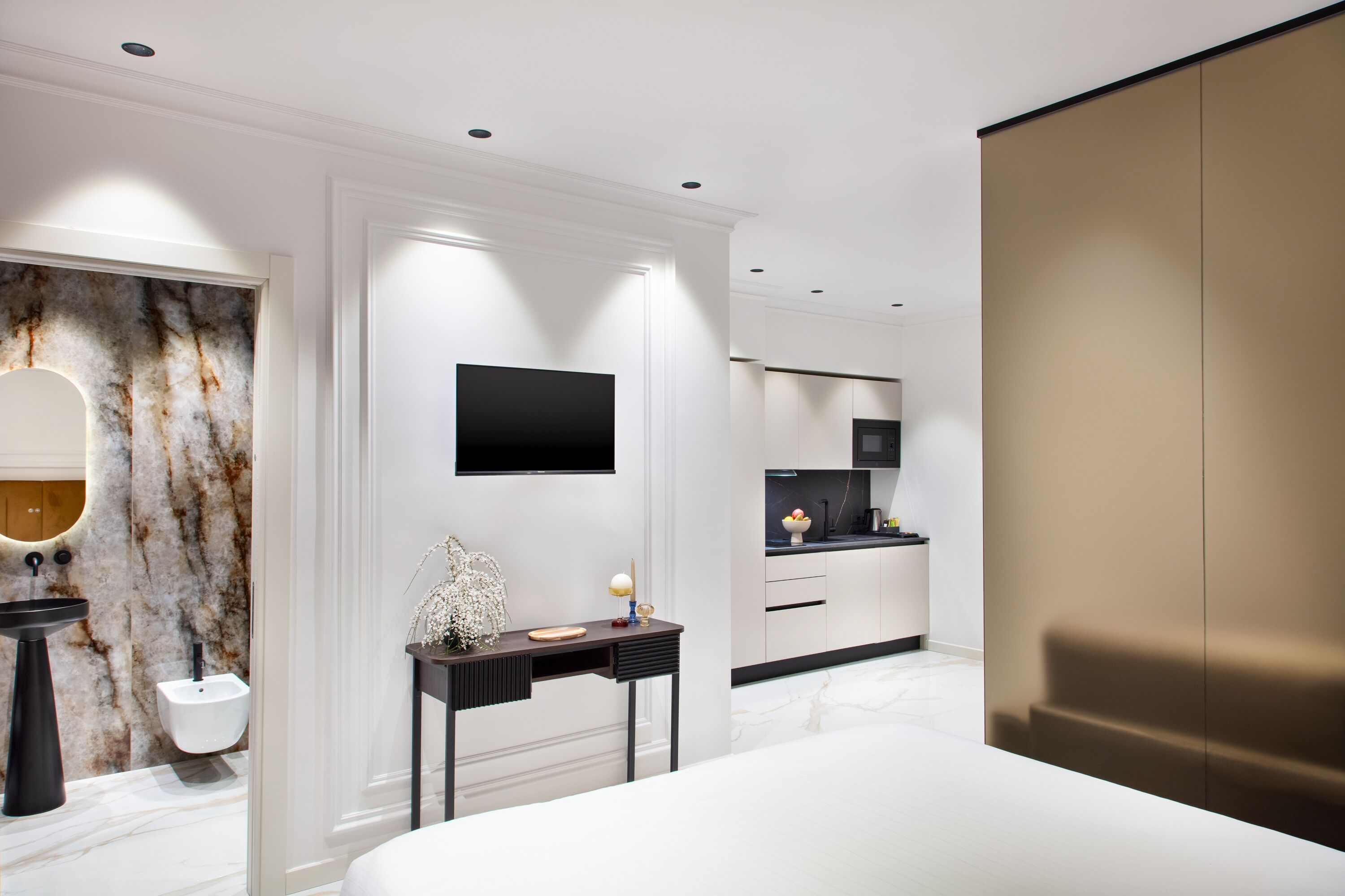 A modern bedroom with a large bed, a bedside table, a flat-screen TV, a bathroom with a marble wall, and a kitchen area.