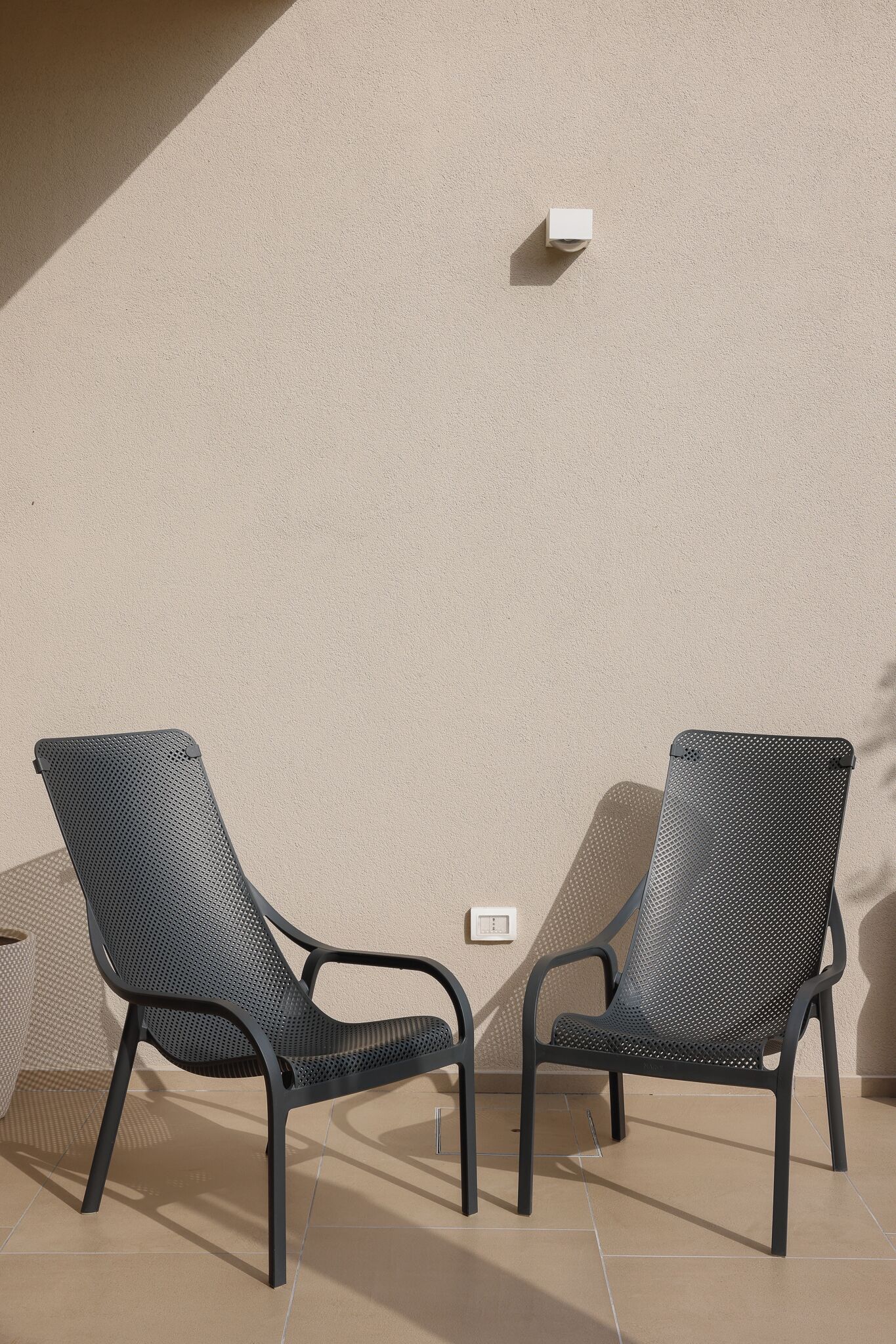 Two black metal chairs with perforated backs and legs, positioned against a beige wall with a small white switch.