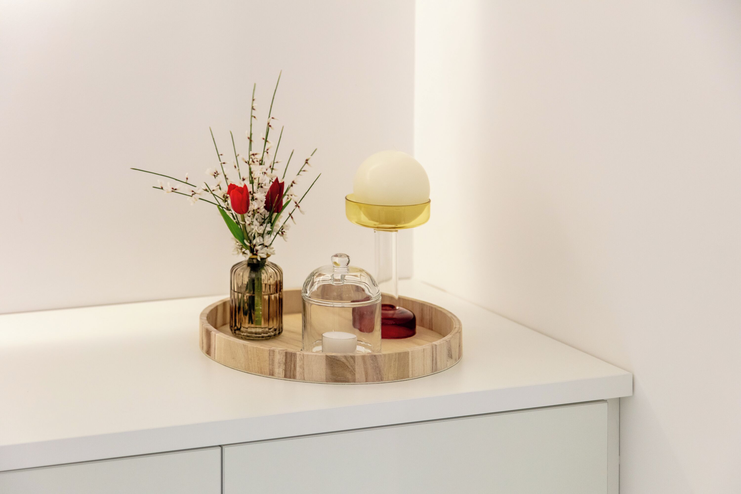 Una encimera blanca con una bandeja de madera que sostiene un jarrón con flores, una vela y un recipiente de vidrio.