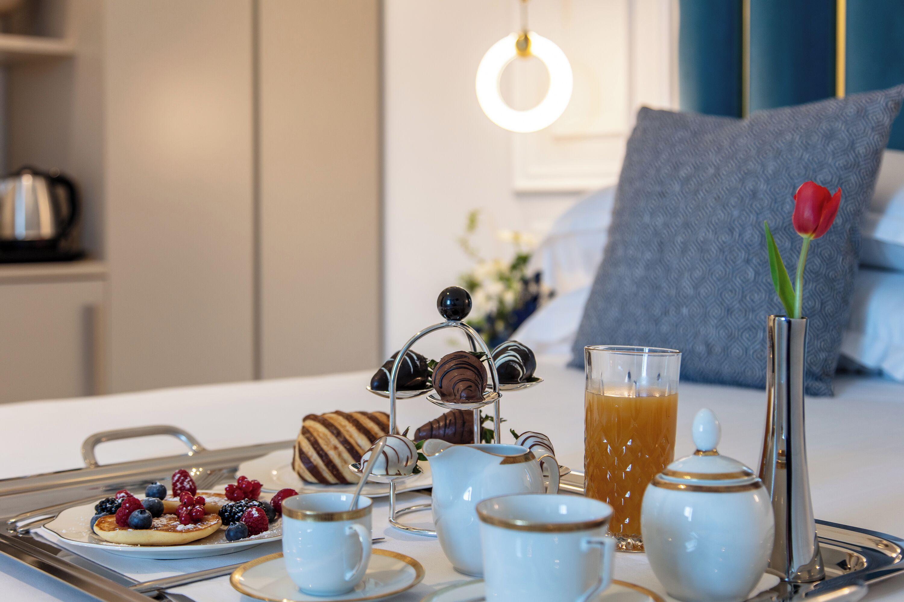 A breakfast tray with pastries, tea, and a vase with a single red tulip on a hotel bed.