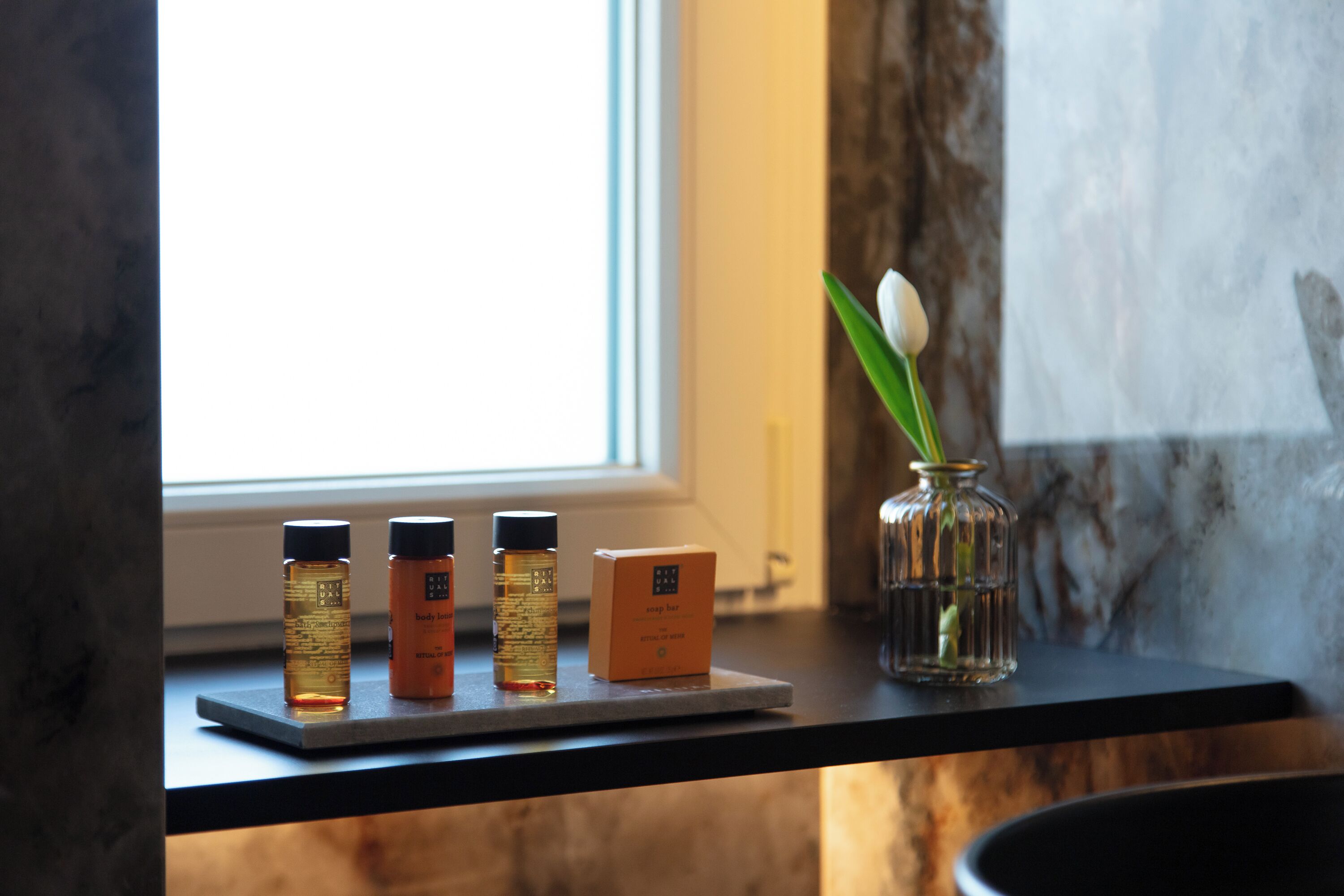 A shelf with three bottles of body lotion and a vase with a single flower.