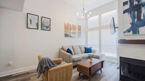 Ein Wohnzimmer mit Sofa, Sessel, Couchtisch und Kamin. An der Wand befinden sich gerahmte Kunstwerke und ein Kronleuchter.