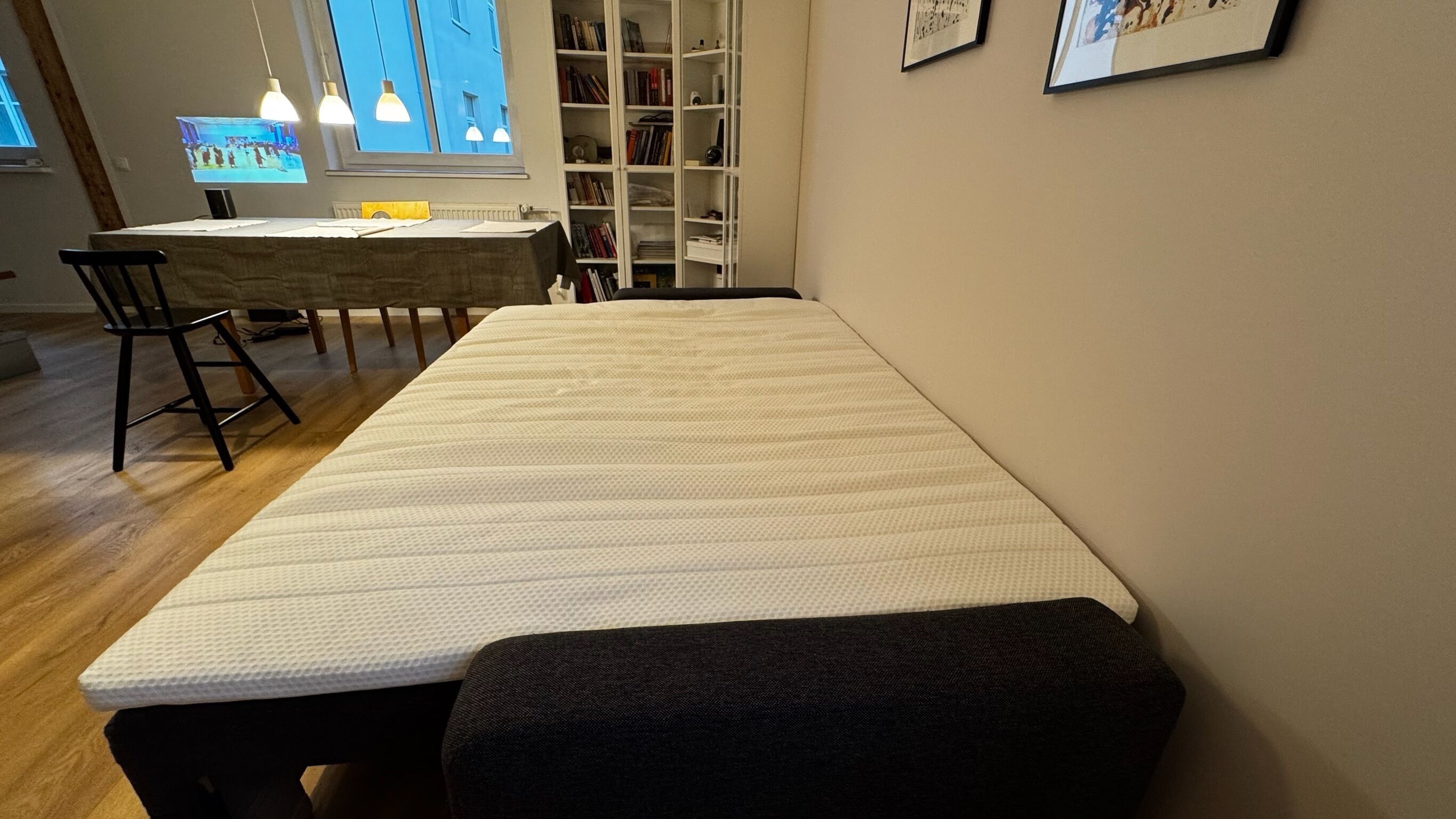 Ein Zimmer mit einem langen, weißen Bett, einer dunklen Bank, einem Holzesstisch mit Stühlen und einem Bücherregal.