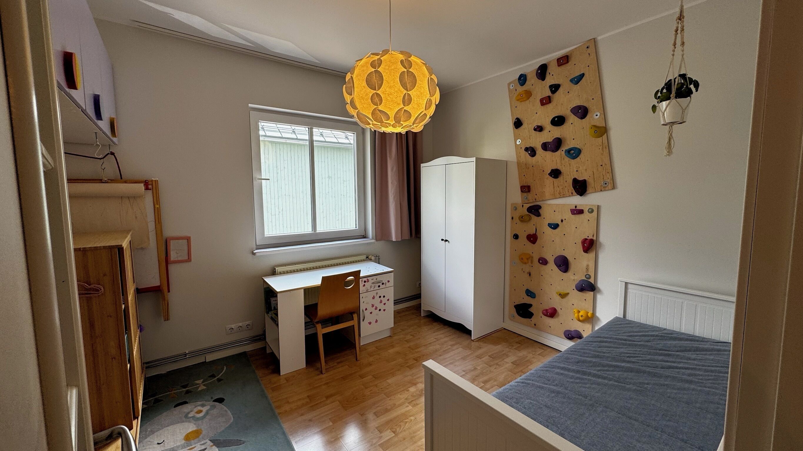Ein Kinderzimmer mit Bett, Schreibtisch, Kleiderschrank, Kletterwand und einer Pendelleuchte.
