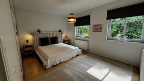 Un dormitorio con cama, mesitas de noche, lámpara colgante, ventana con vista a follaje y radiador.