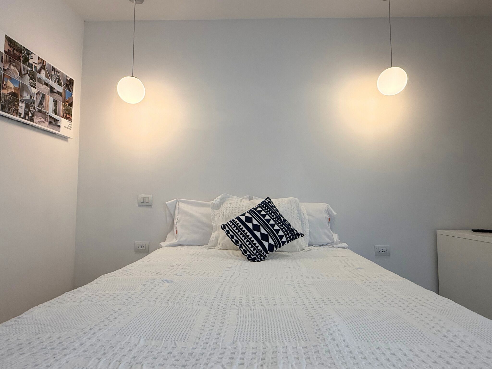 A neatly made bed with a white duvet and a patterned pillow, two pendant lights hanging above, and a wall-mounted shelf with a photo collage.
