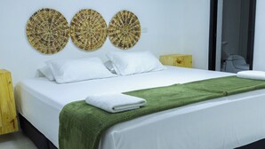 Un lit bien fait, avec du linge de lit blanc, un plaid vert et une serviette blanche. Trois éléments décoratifs en fibres tressées sont accrochés au mur au-dessus du lit.