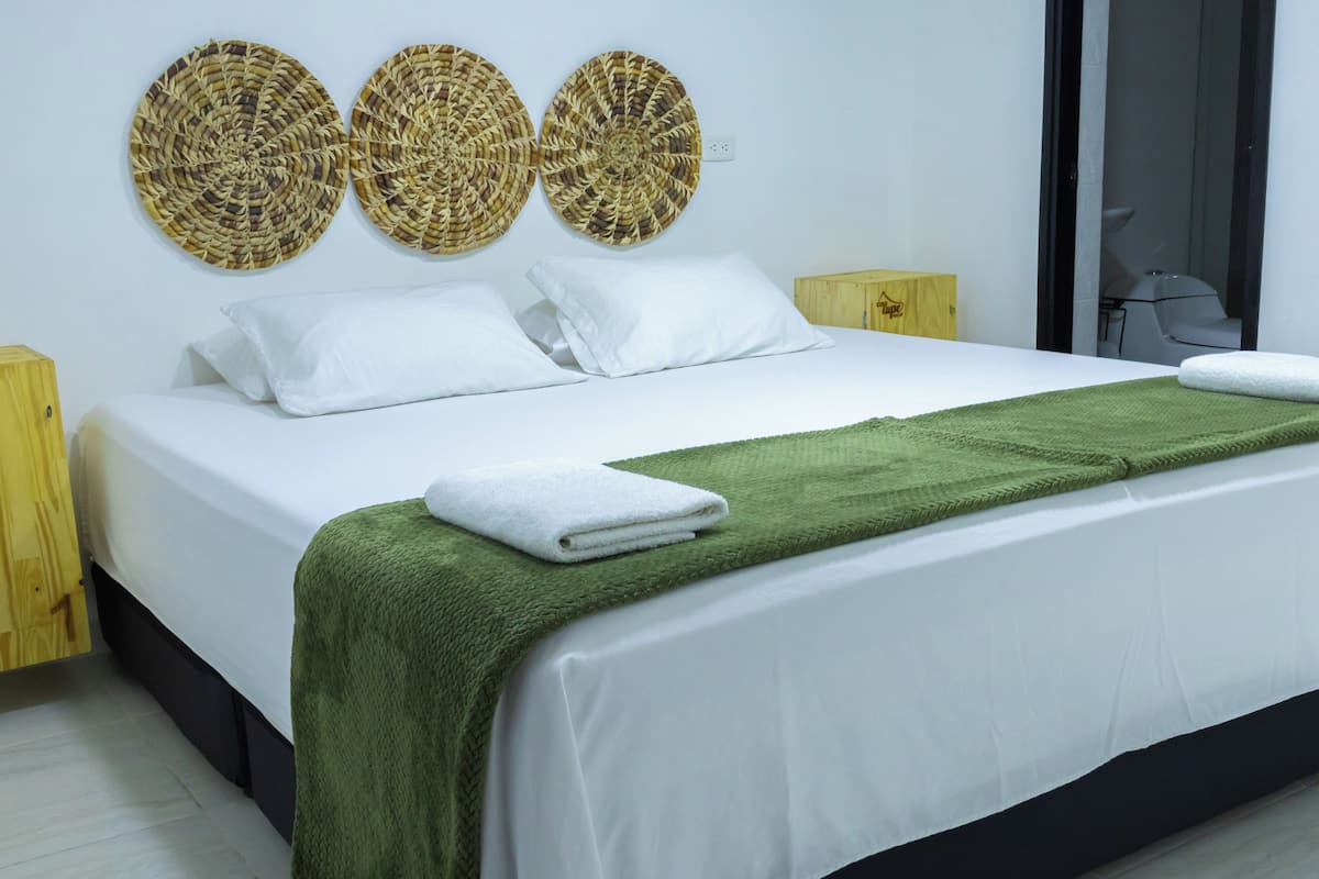 Un lit bien fait, avec du linge de lit blanc, un plaid vert et une serviette blanche. Trois éléments décoratifs en fibres tressées sont accrochés au mur au-dessus du lit.