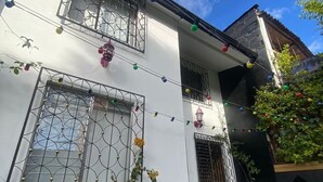 Une maison à deux étages, avec une façade blanche, des moulures noires et un balcon. Elle possède deux fenêtres avec des barreaux en métal et des guirlandes lumineuses au-dessus.