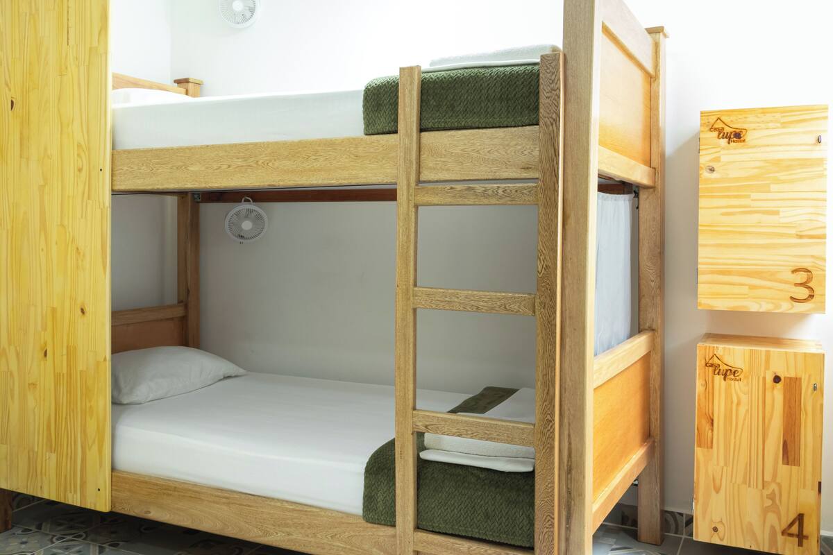 Un lit superposé avec une échelle, un ventilateur et une petite armoire en bois portant les numéros 3 et 4.