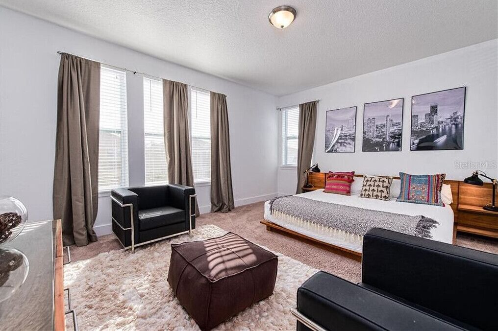 Une chambre à coucher avec un lit, un canapé noir, un pouf marron, une table basse en verre et une décoration murale représentant un paysage urbain.