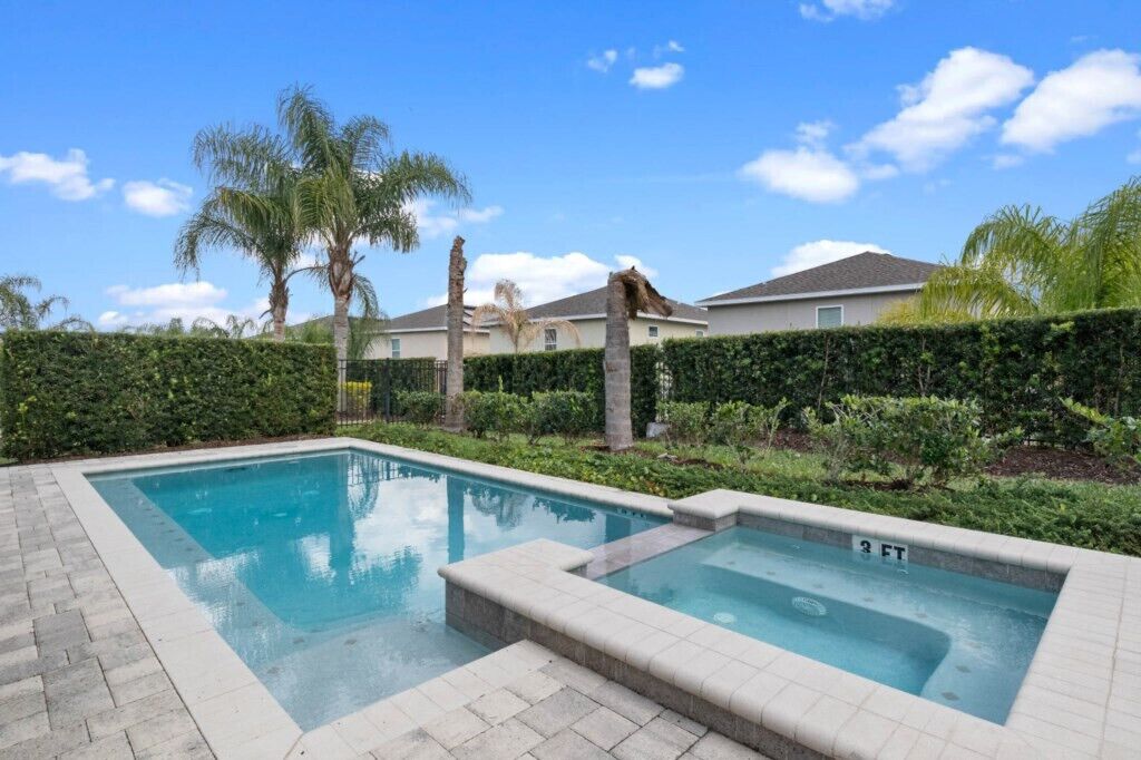 Una piscina rectangular con un jacuzzi, rodeada por un seto y palmeras, con casas al fondo.