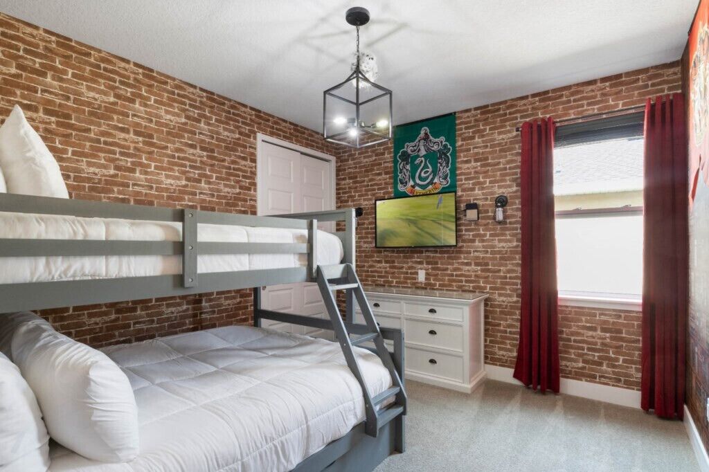 A room with a bunk bed, a desk, a chair, a white dresser, a red curtain, a window, and a wall-mounted light fixture.