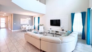 A modern living room with a sectional sofa, a flat-screen TV, and a dining area with a white table and chairs.
