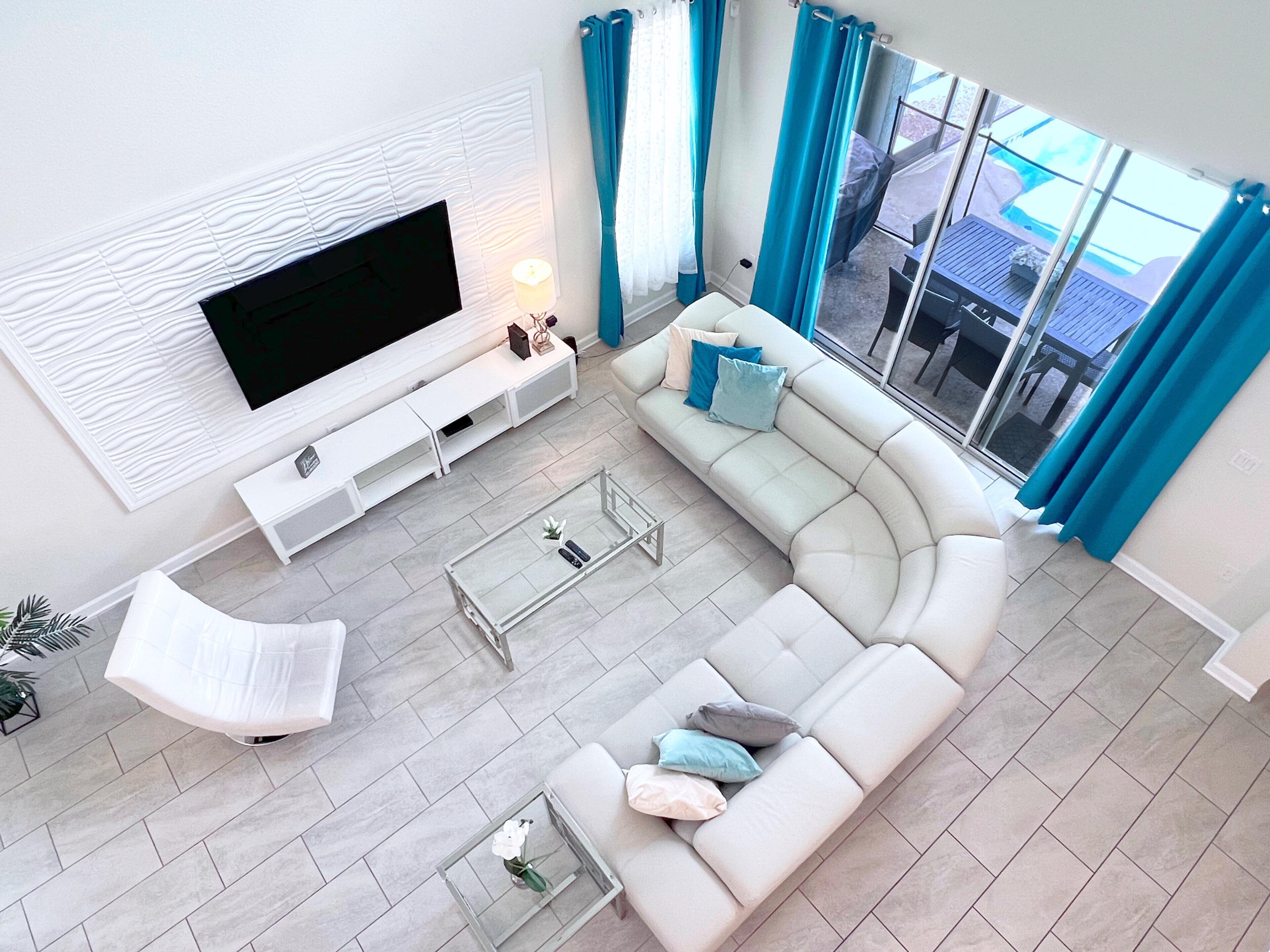 Ein modernes Wohnzimmer mit einem großen Flachbildfernseher, einem weißen Sofa, einem Glastisch und Blick ins Freie durch große Fenster.