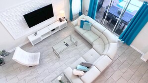 A modern living room with a large flat-screen TV, a white sofa, a glass coffee table, and a view of the outdoors through large windows.