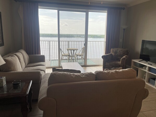 A living room with a sofa, armchair, and a TV on a stand. There is a balcony with a view of the water.