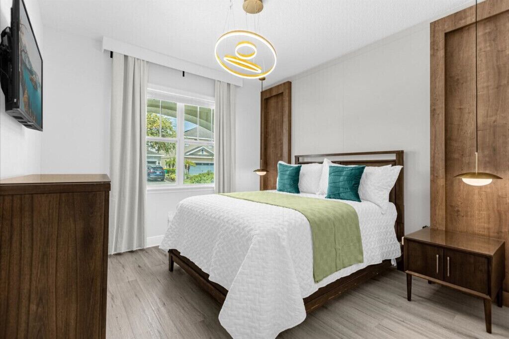 A bedroom with a bed, a bedside table, a window with curtains, and a wall-mounted light fixture.
