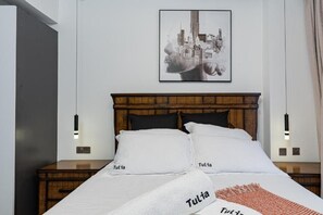 A neatly made bed with white bedding and pillows labeled "Tulía", a framed cityscape artwork on the wall, and a wooden nightstand with a lamp.