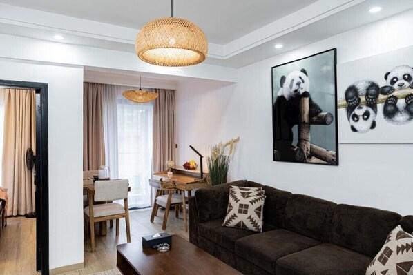 A modern living room with a dark brown sofa, a wooden coffee table, and a dining area with a wooden table and chairs. There is a large panda artwork on the wall.