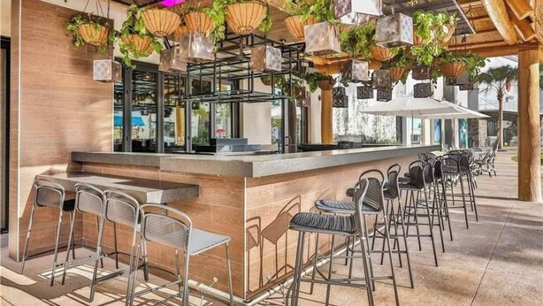 Ein Außenbereich mit Bar, ausgestattet mit Metallstühlen und -tischen, hängenden Pflanzgefäßen und einer Holzbalkendecke.