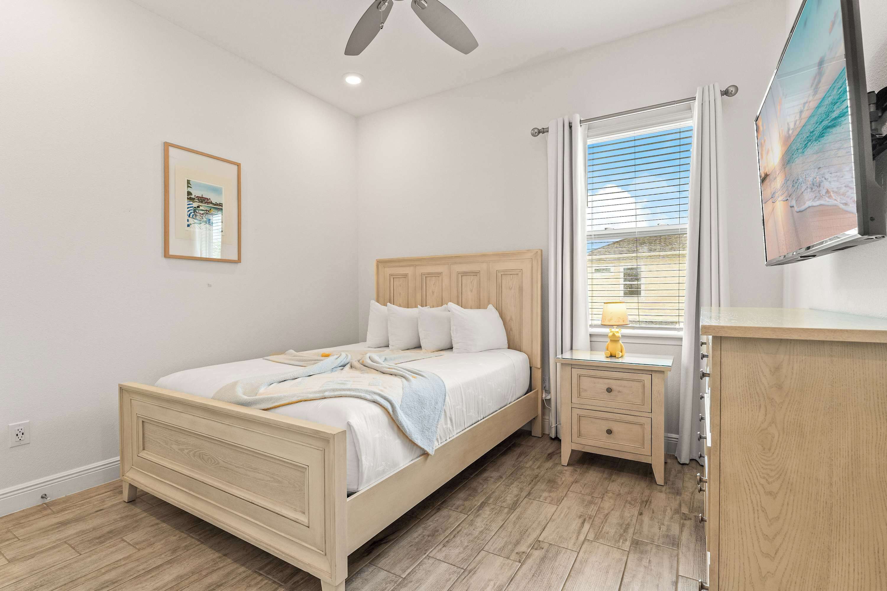 A bedroom with a wooden bed, a nightstand, a dresser, a ceiling fan, a window with curtains, and a framed picture on the wall.