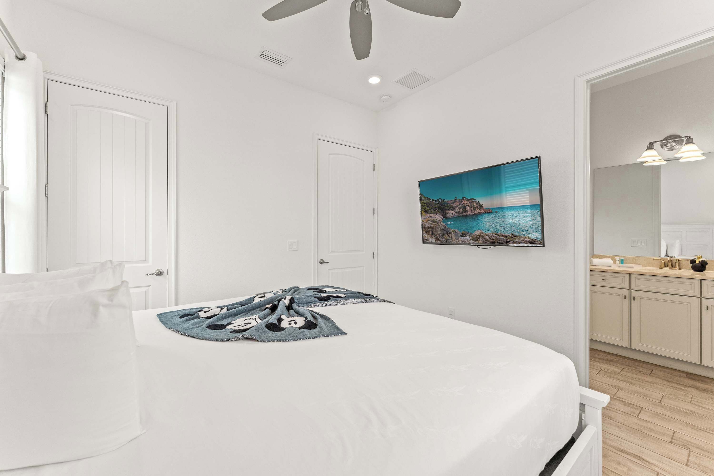 A bedroom with a bed, a ceiling fan, a painting of a coastal scene, and a bathroom with a sink and mirror.