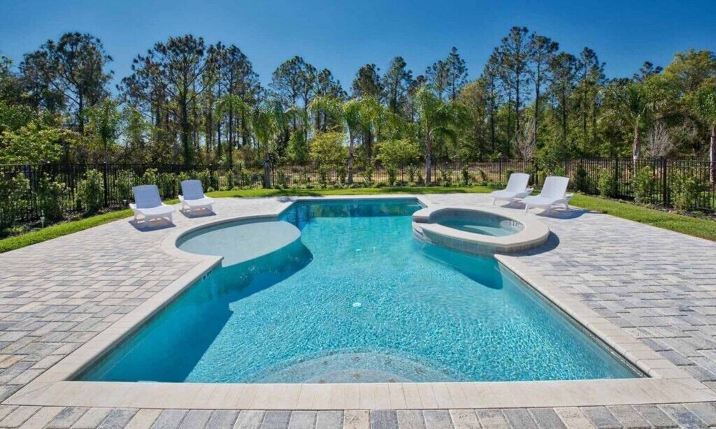 A rectangular swimming pool with two circular hot tubs, surrounded by a paved deck and a fence.