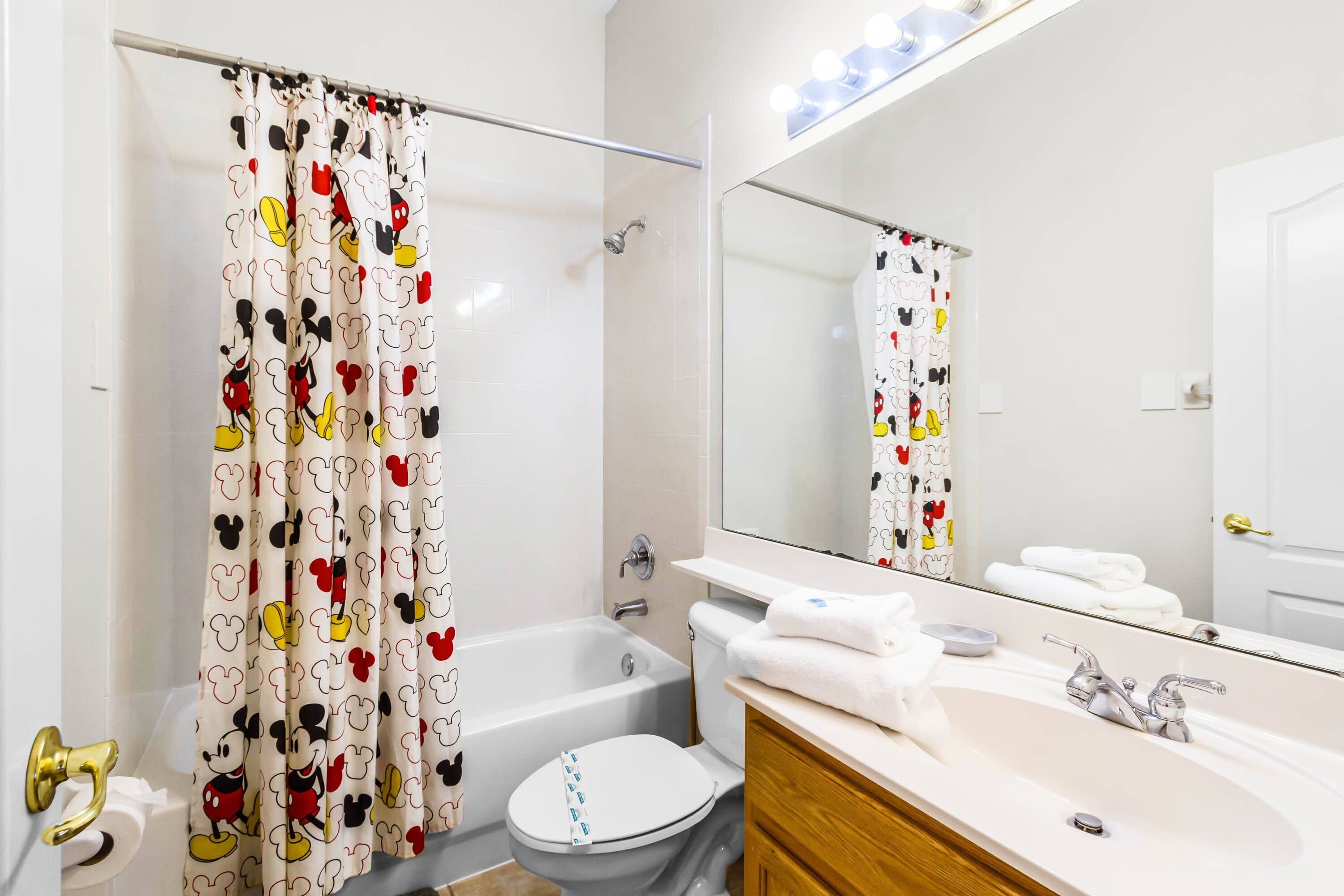 A bathroom with a Mickey Mouse themed shower curtain, a bathtub, a toilet, a sink with a mirror, and a towel rack.