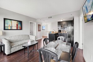 A modern living room with a sofa, dining table, and chairs. A kitchen is visible in the background with cabinets and appliances.
