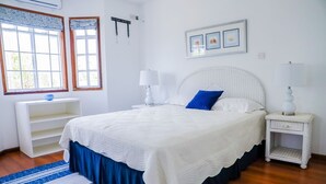 A neatly arranged bedroom with a large bed, bedside tables, a window with curtains, and a wall-mounted shelf.
