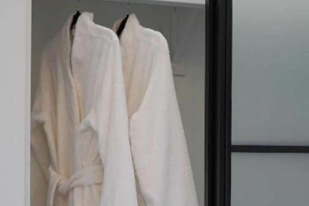 Two white bathrobes hanging on a rack in a room with a plain wall and a black-framed mirror.