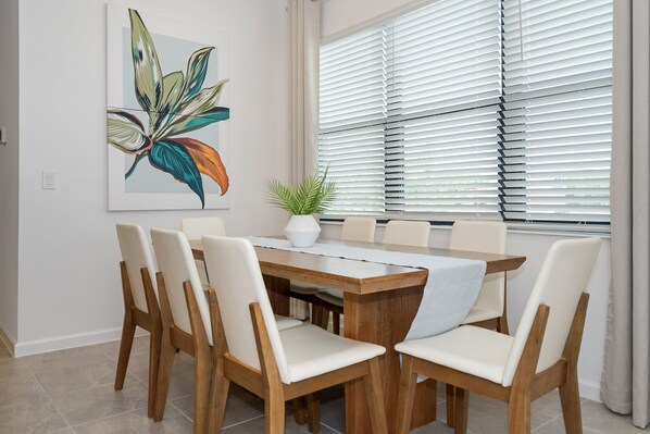 A dining area with a wooden table and chairs, a potted plant, and a large window with blinds.