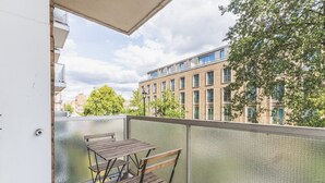 A balcony with a wooden table and chairs, a glass railing, and a view of a building with multiple windows and balconies. - Belgravia 2 BR Hideaway with Balcony (London)