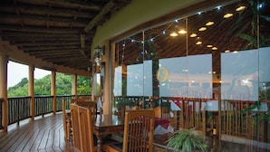 A wooden deck with tables and chairs, large windows with reflections, and a view of greenery. - Mara Engai Wilderness Lodge (Nairobi)