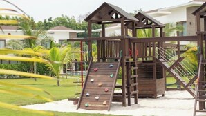 Ein Spielplatz mit Rutsche und Klettergerüsten, umgeben von einer Rasenfläche und Palmen.
