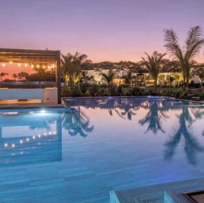 Vista a la piscina al atardecer, con un edificio moderno, palmeras y cielo despejado.