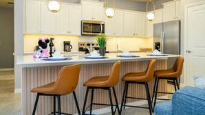 A modern kitchen with white cabinets, a stainless steel refrigerator, a microwave, and a coffee maker. There are three brown bar stools and a small island with a plant.