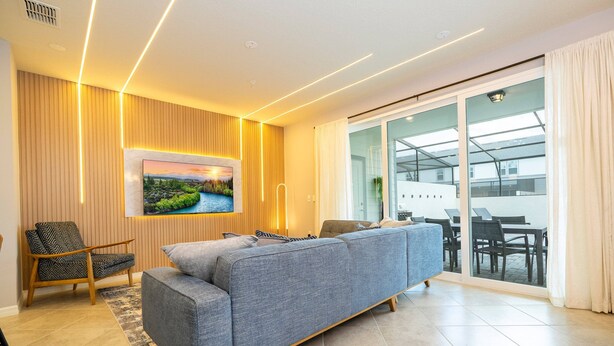 A modern living room with a grey sofa, wooden panel walls, a large TV, and a glass door leading to a balcony.
