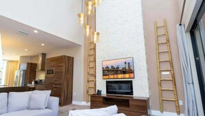 A spacious living room with a high ceiling, a large flat-screen TV mounted on a white brick wall, and a modern chandelier.