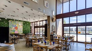 Un restaurant avec de hauts plafonds, de grandes fenêtres et un mur décoré d’une fresque végétale. Les tables sont dressées avec de la verrerie bleue et des chaises en bois.
