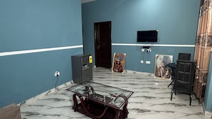 Un soggiorno con pareti color petrolio, una ventola a soffitto, una TV a muro, un tavolino da caffè in vetro, un divano e un tappeto decorato.
