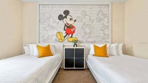 A hotel room with two beds, white bedding, yellow pillows, and a large Mickey Mouse mural on the wall.