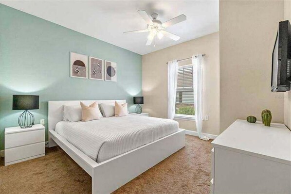 Un dormitorio con cama, mesitas de noche, ventilador de techo, ventana con persianas y televisor sobre un mueble.